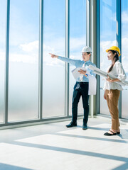 Asian architect Male and female inspecting construction work inside high-rise building near huge window glass on sky background.