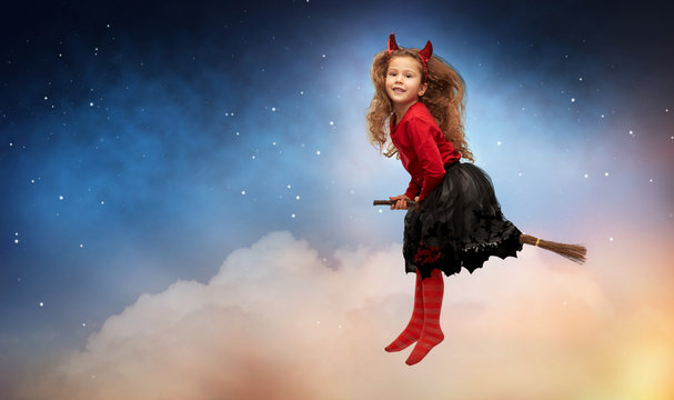 halloween, holiday and childhood concept - smiling girl in party costume and red devil's horns flying with witch's broom over stars in night sky on background