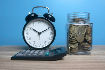 Selective focus of vintage alarm clock,calculator and a jar full with gold coins on wooden table with a blue background.