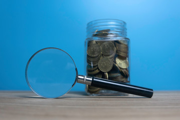 Selective focus of jar full of gold coins and magnifying glass on wooden table with a blue background.