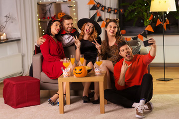 friendship, holiday and people concept - group of happy smiling friends in halloween costumes of vampire, devil, witch and cheetah taking selfie by smartphone at home party at night