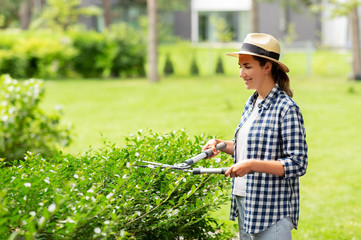 gardening and people concept - happy smiling young woman in straw hat with pruner or pruning shears cutting branches at summer garden