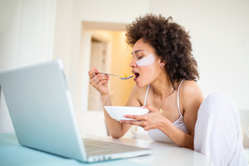 Attractive woman using her laptop computer while having breakfast and studying in home indoors. Freelance female model girl working on her laptop in the morning . Cup of milk with corn flakes