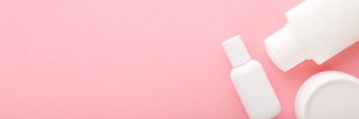 White cream bottles and jar on light pink background. Pastel color. Care about face, hands, legs and body skin. Beauty products for women. Empty place for text or logo. Wide banner. Top down view.