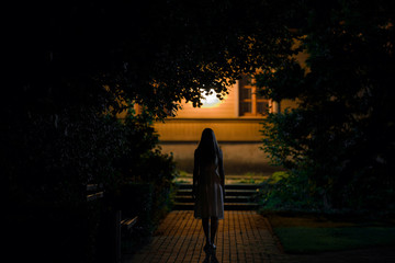 Young alone woman in dress walking on sidewalk through dark park to home in summer black night....