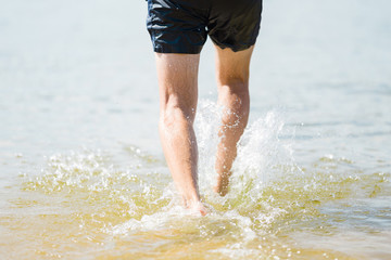 Young man legs walking in sea water in sunny day. Back view. Closeup.