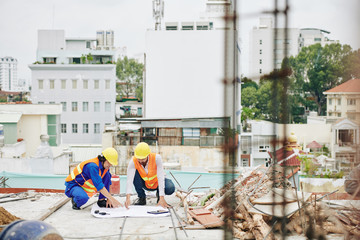 Builders in orange vests and hardhats pointing at building plan on the floor and discussing plan of...