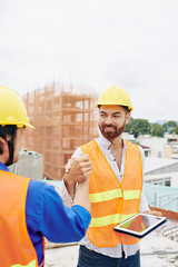 Cheerful bearded contractor with tablet computer shaking hand of coworker when greeting him in the morning