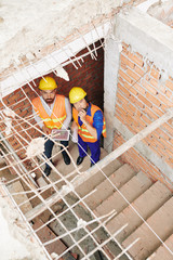 Builders with tablet computer stading on stairs in building under construction, talking on...