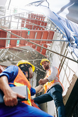 Builder in vest and hardhat helping coworker to climb up the scaffolding outside the building under...