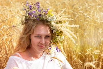 Beautiful blonde with long hair and a wreath of flowers on her head sits in a field of ripe wheat.