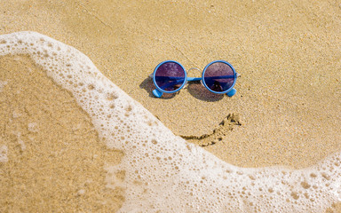 Fototapeta na wymiar Sunglasses lie on the beach next to the sea. A smile is drawn under the glasses. The wave washes away the smile.
