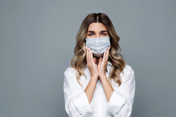 Young sad woman with curly hair dressed in white shirt looking at camera, wearing protective mask against covid-19, feels symptoms of a cold, keeps hands near head, isolated on grey background. 