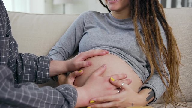 Front view of charming smiling happy woman with dreadlocks that sitting on the couch and enjoying when her husband touching with hands on her pregnant tummy
