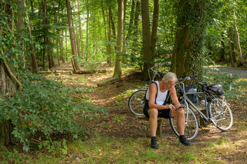 Senior man in cycling shorts sitting on a bench in a forest staring at his phone. There are a few bicycles parked against the bench.
