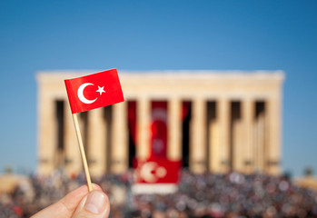 Male hand holding a mini Turkey flag in front of Anitkabir and blurred crowd.. August 30 Zafer...