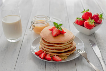 Homemade fresh pancakes with berries, honey and milk on a white wooden background. Rustic, American traditional breakfast.