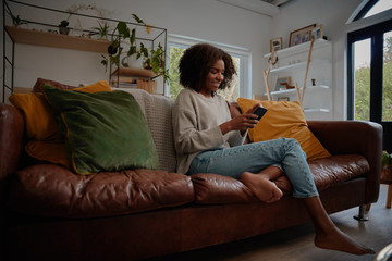 Smiling african woman sitting on comfortable couch using smartphone at home during lockdown