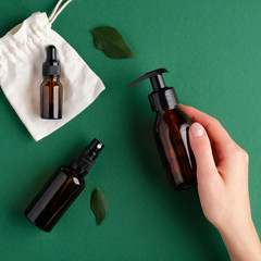 Female hand holding pump amber glass bottle over green background with spray bottle and essential oil dropper bottle. SPA natural organic beauty products set. Flat lay, top view