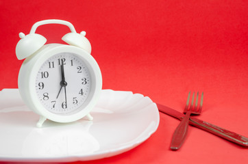 white alarm clock sits on a white plate on a red background with a fork and knife close up. concept time for dinner, creative