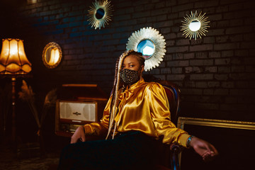 African American fashionable woman wearing stylish dark green velvet protective face mask with golden rhinestones, yellow blouse, posing in dark vintage luxury interior. Copy, empty space for text