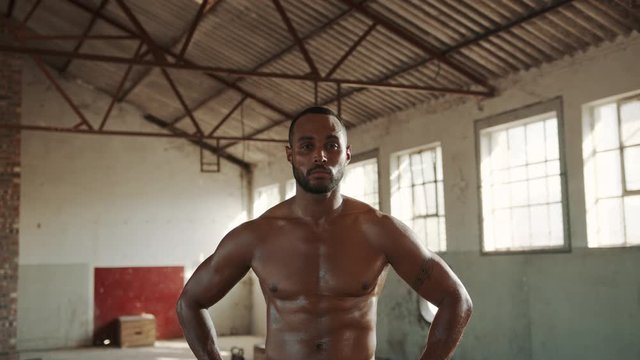 Fit man standing shirtless inside abandoned warehouse with his hands on hips. Strong man in fitness studio looking at camera.

