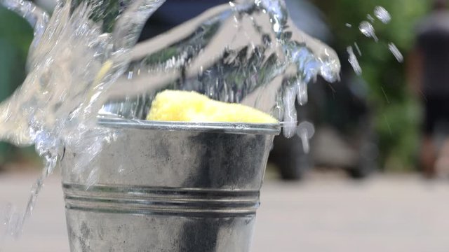 A man washes a car on the street, then throws a sponge into a distant bucket with splash of detergent. A domestic hand washing car with soapy water.