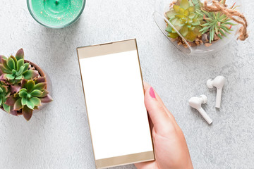 Woman holding smartphone with blank screen on concrete desk with wireless earphones, succulent...