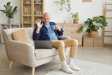 Obraz premium Full length portrait of modern senior man waving at camera and smiling happily during video call at home, copy space