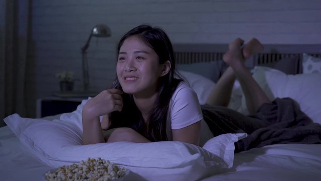closeup asian young woman slacking off in dark bedroom watching hilarious talk show at night. girl in self-quarantine eating popcorn and laughing happily at  joke told by tv host. genuine lifestyle