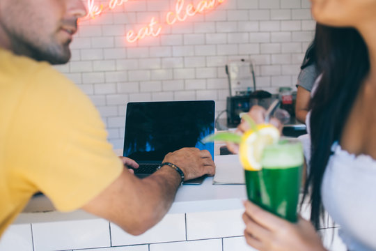 Cropped image of male typing on laptop computer while female holding glass with fresh smoothie spending time in cafe interior, rear view of young woman and man searching information browsing web page