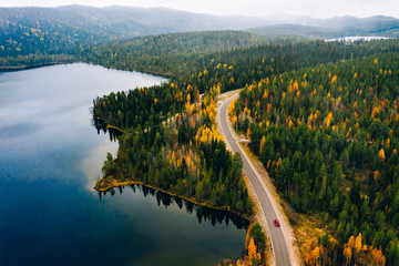 Aerial view of rural road with red car in yellow and orange autumn forest with blue lake