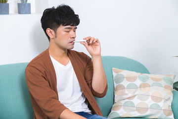 Sad unhappy handsome man sitting on the sofa and using ear thermometer while having headache and...