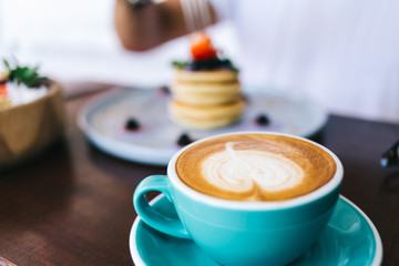 Cropped image of blue coffee cup with cappuccino hot aroma caffeine beverage with foam standing on table on blurred background with tasty delicious pancakes meal on breakfast in cafeteria