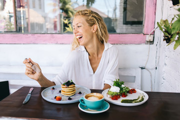 Obraz na płótnie Canvas Cheerful caucasian female sitting at cafe interior on breakfast meal laughing eating tasty salads and pancakes, happy hipster girl satisfied with delicious healthy diet nutrition keeping lifestyle