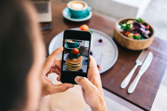 Cropped image of male influencer making photo of food for social networks via smartphone camera. guy blogger holding mobile phone taking picture of flatlay tasty pancakes stack on brunch in cafe