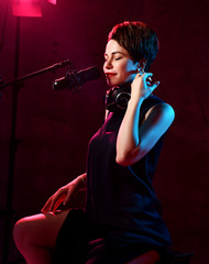 Inspired short haired brunette woman in elegant black dress sits in studio in headphones and sings in microphone