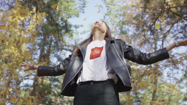 Brown-haired woman in shirt and black jacker standing in autumn forest under falling yellow leaves and laughing joyfully. Low angle 4K 360 degree tracking arc shot.