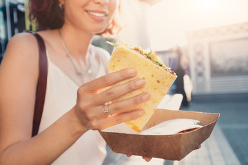 Happy woman eats a fast food flatbread with filling sitting on the street and resting after a working day. Concept of healthy food and extra calories