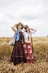 Fototapeta premium Two hippie women, wearing boho style clothes, dancing in the wheat field, having fun. Female friends, traveling together in countryside. Eco tourism concept. Summer leisure free time entertainment