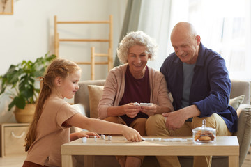 Warm toned portrait of happy senior couple playing board games with cute little granddaughter at...