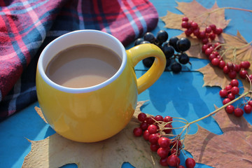 Yellow cup of coffee, plaid, red berries, and autumn leaves on blue wooden background.  Cozy autumn atmosphere. Copy space, selective focus