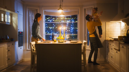 Happy Family of Three Cooking and Having Dinner Together. Mother Prepares Food, Father Hugs Cute Little Girl. Festive Table in Cozy Kitchen