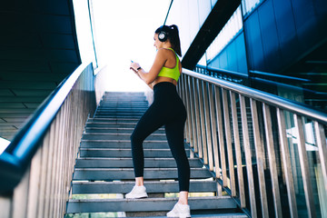 Back view of sportive female bodybuilder with perfect muscular body shape checking fit results on smartphone device standing at urban stairs, sport woman watching streams with web advices for workout