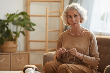 Warm toned portrait of elegant senior woman knitting and looking at camera while sitting on couch...
