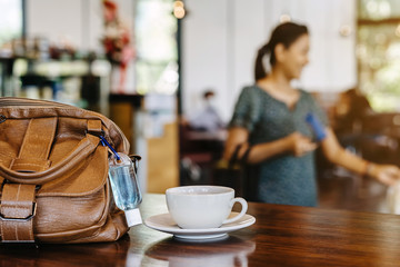 Mini alcohol gel bottle to kill Corona Virus(Covid-19) hang on a brown leather shoulder bag with hot coffee on table in cafe.New normal lifestyle.Health care concept.Selective focus on alcohol gel