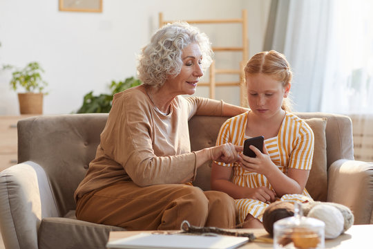 Warm toned portrait of elegant senior woman showing smartphone photos to cute granddaughter while sitting on couch in cozy home lit by sunlight