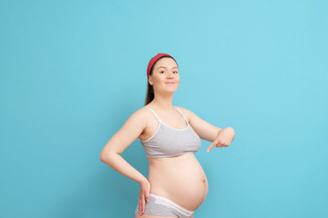 happy young pregnant woman on blue background, woman points finger to belly