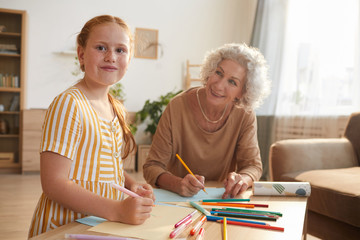Fototapeta premium Warm toned portrait of cute red haired girl smiling cheerfully at camera while drawing together with grandma enjoying time at home, copy space