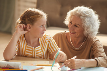 Warm toned portrait of smiling senior woman looking at cute red haired girl and drawing together...